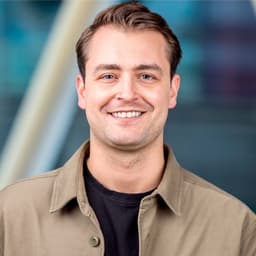 Ein Mann mit einer beigen Jacke mit Kragen. Er trägt einen Drei-Tage-Bart, hat kurze Haare und lächelt.