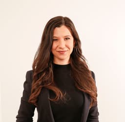Eine Frau mit braunen, langen und welligen Haaren. Sie trägt einen schwarzen Blazer.