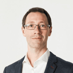 Ein Mann mit kurzen, dunklen Haaren. Er trägt eine Brille und ist in einem Sakko und Hemd gekleidet.