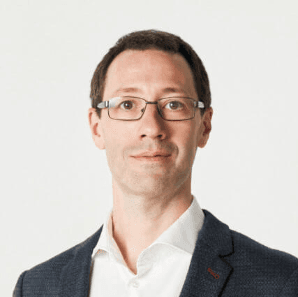 Ein Mann mit kurzen, dunklen Haaren. Er trägt eine Brille und ist in einem Sakko und Hemd gekleidet.