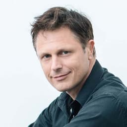Ein Mann mit kurzen, dunkelbraunen Haaren. Er lächelt und trägt ein dunkelblaues Hemd.