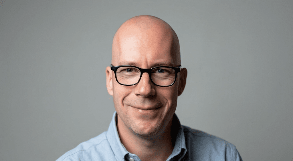 Ein Mann mit einer Glatze und einer schwarzen Brille. Er trägt ein hellblaues Hemd und lacht.