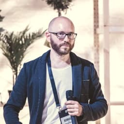 Ein Mann mit einer schwarzen Brille und Bart. Er trägt eine Glatze und ist Business Casual gekleidet.