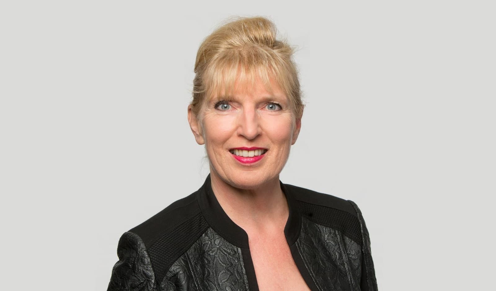 Eine Frau mit blonden Haaren. Sie trägt einen schwarzen Blazer, der leicht schimmert.