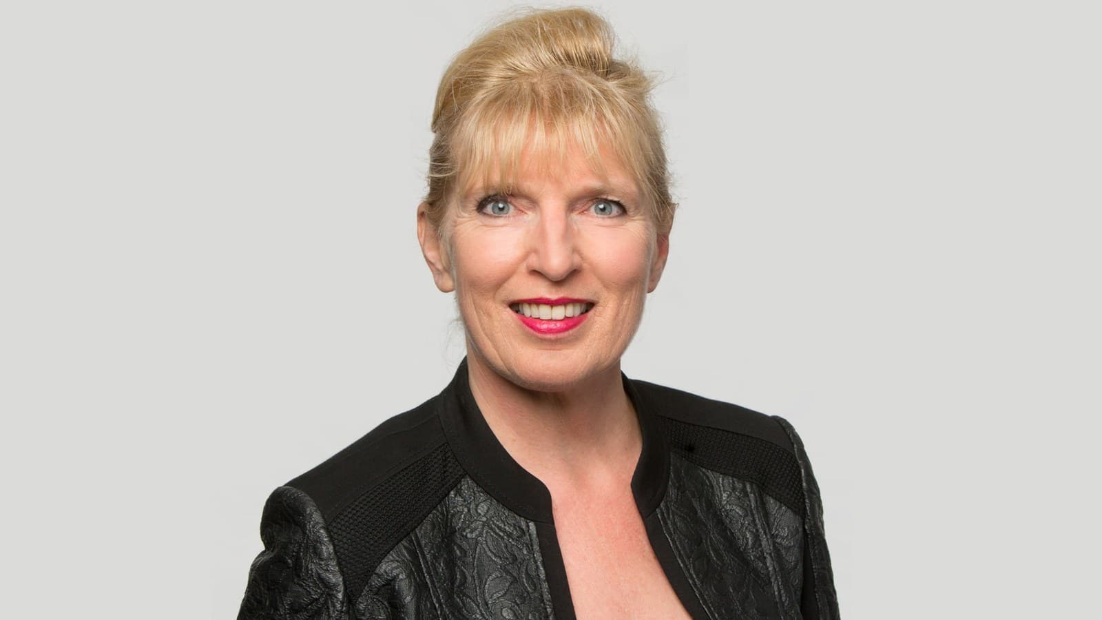 Eine lachende Frau mit blonden Haaren und blauen Augen, gekleidet in einem schwarzen Blazer